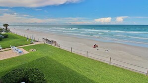 A beach view with a grassy area, a staircase leading down to the beach, and a fence along the shore. - Colony Beach Club 223 (New Smyrna Beach)