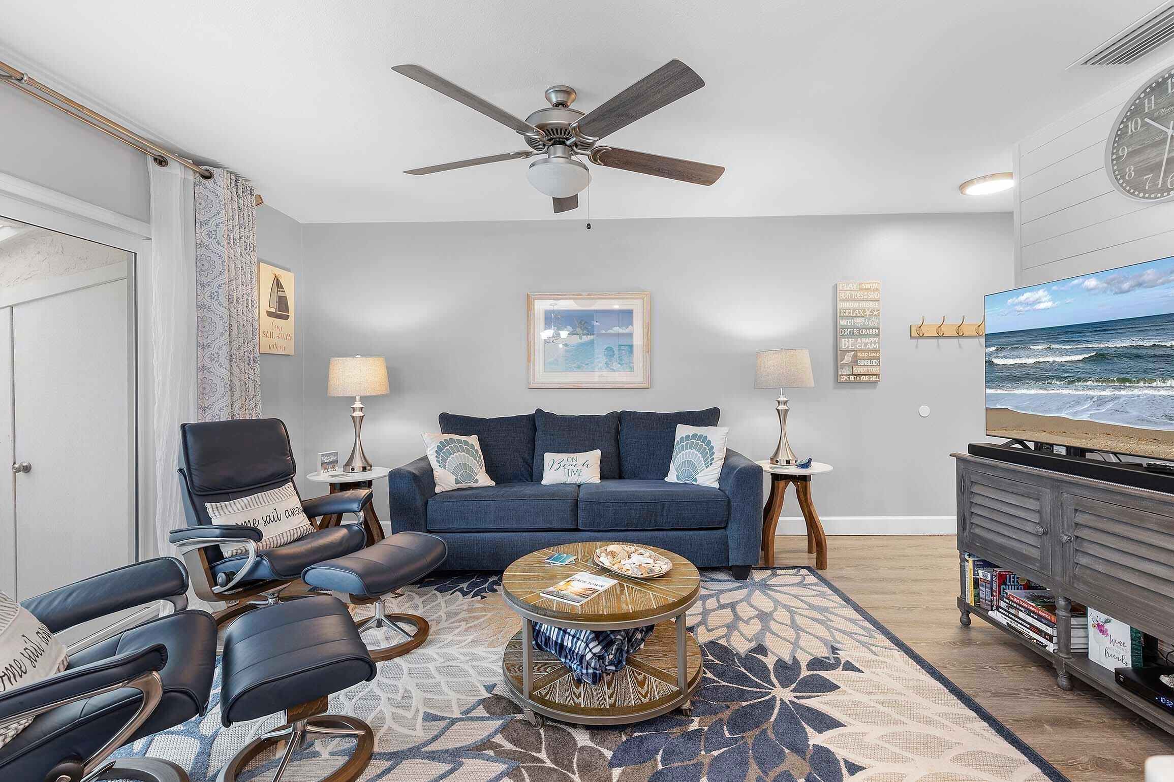 A modern living room with a blue sofa, a round coffee table, a ceiling fan, and a television on a wooden stand.