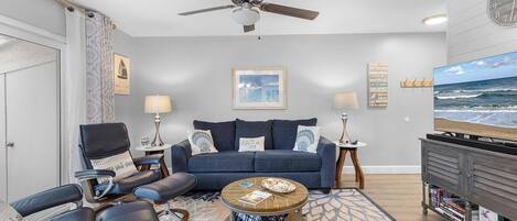 A modern living room with a blue sofa, a round coffee table, a ceiling fan, and a television on a wooden stand.
