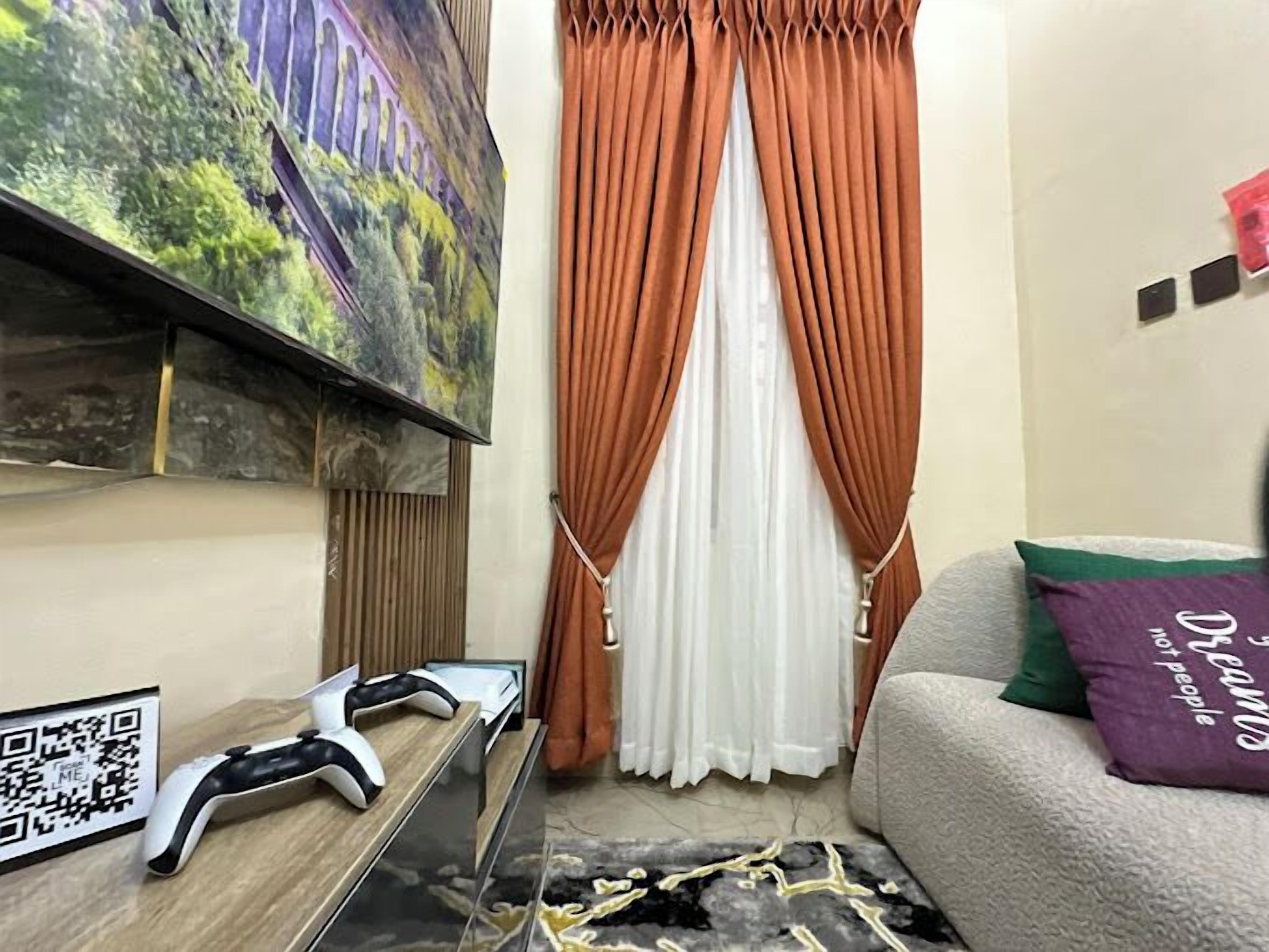 A living room with a flat-screen TV mounted on the wall, a gaming console on a wooden table, a couch with a purple pillow, and a window with orange curtains.