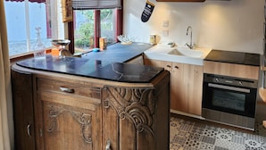 Een keuken met een centraal eiland, houten kastjes, een inbouwoven en een raam met jaloezieën.