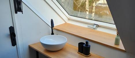 A skylight above a wooden vanity with a white basin, a black faucet, and a small shelf with two black containers.