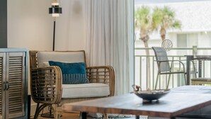 Una habitación con una silla de mimbre, un sofá blanco con un cojín azul, una mesa con un tazón y vistas a palmeras afuera.