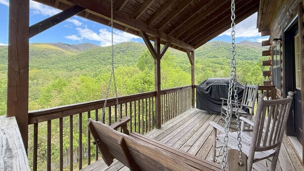 A wooden deck with a swing chair, a covered grill area, and a view of green hills. - Sleepy Bear Log Cabin with View, WiFI (Maggie Valley)