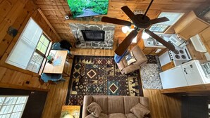 A cabin interior with a stone fireplace, a sofa, a television, a ceiling fan, and a kitchen area. - Sleepy Bear Log Cabin with View, WiFI (Maggie Valley)