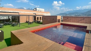 A rooftop pool area with a wooden deck, a small pool, and a seating area with tables and chairs.