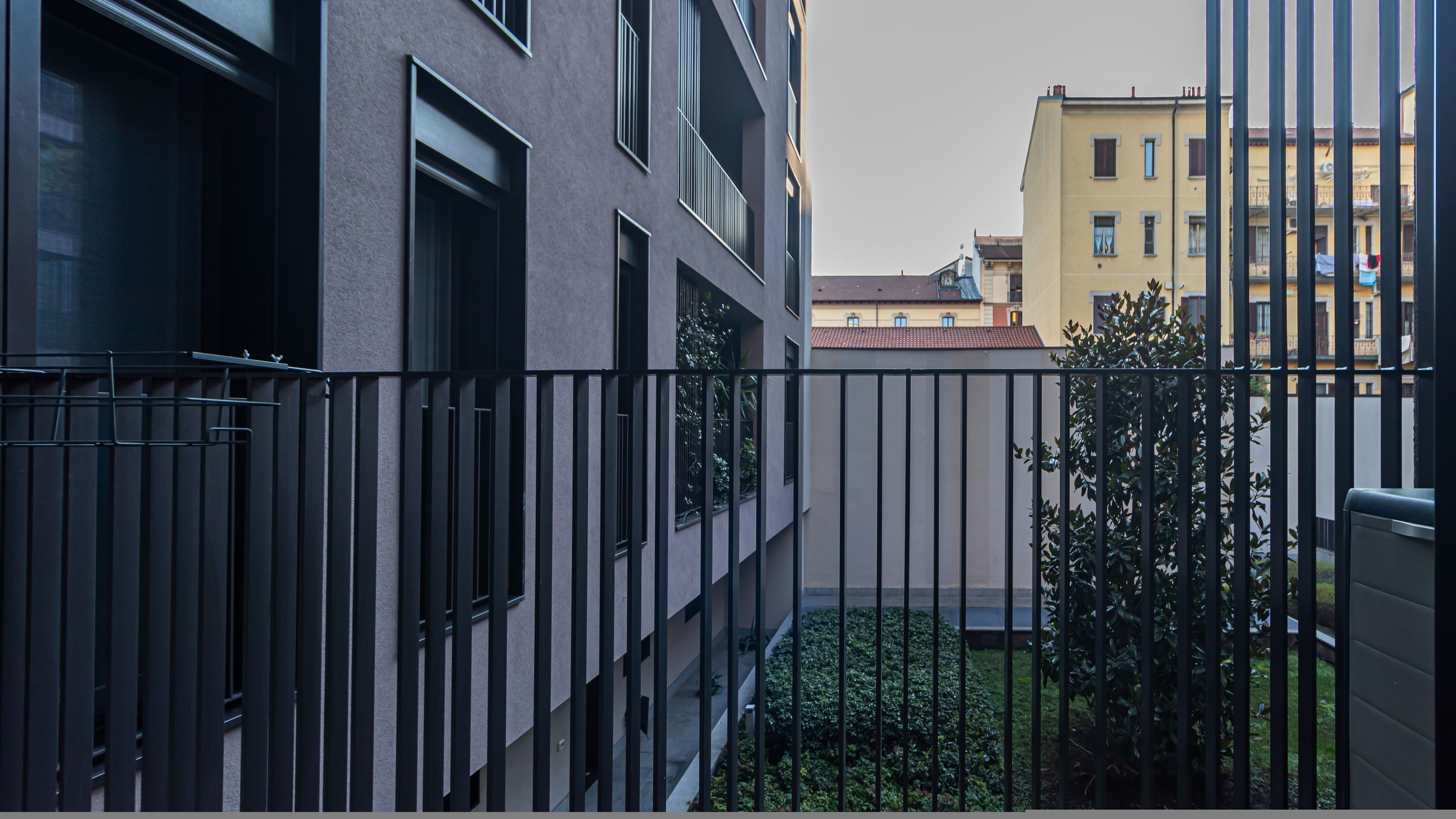 A modern apartment building with a balcony, a metal fence, and a small garden area.