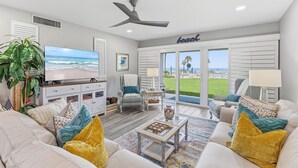 A living room with a TV, seating, a ceiling fan, and a view of the beach.