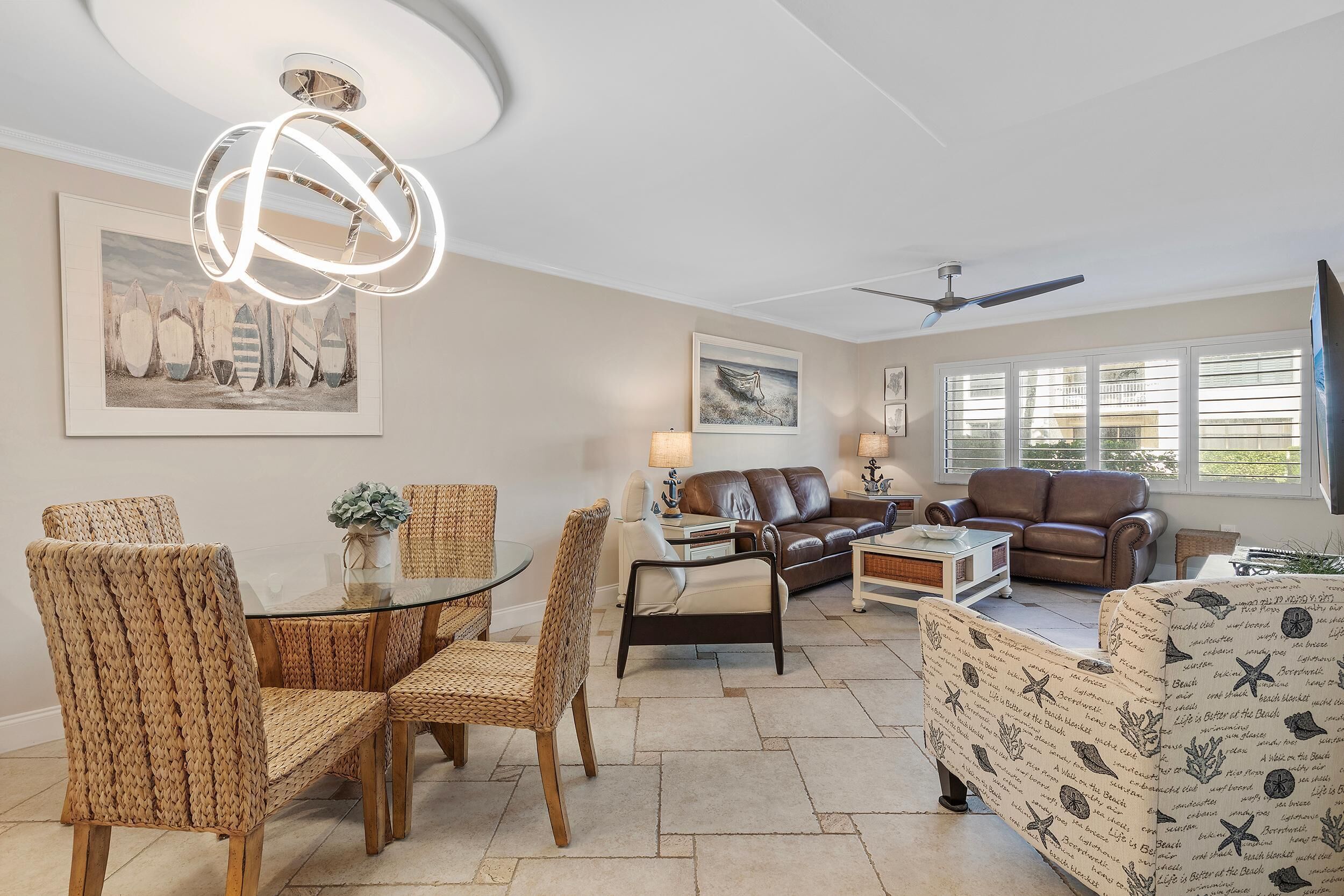 A living room with wicker chairs, a glass table, a brown leather sofa, and a patterned armchair. There is a ceiling fan, a framed picture on the wall, and a window with blinds.