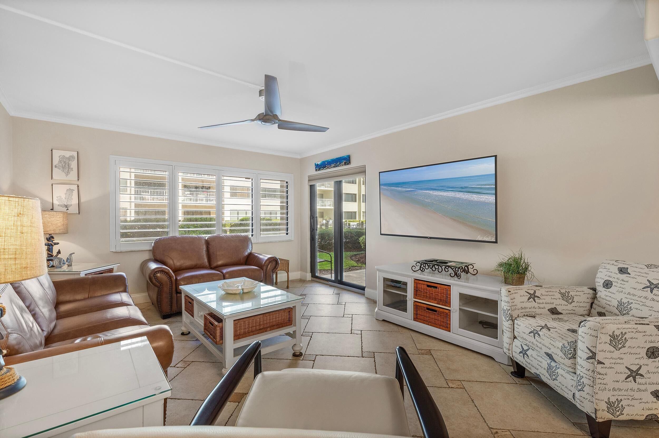 Een ruime woonkamer met een plafondventilator, een groot raam met jaloezieën, een bruine leren bank, een witte fauteuil, een glazen salontafel en een groot gepresenteerd foto van een strand.