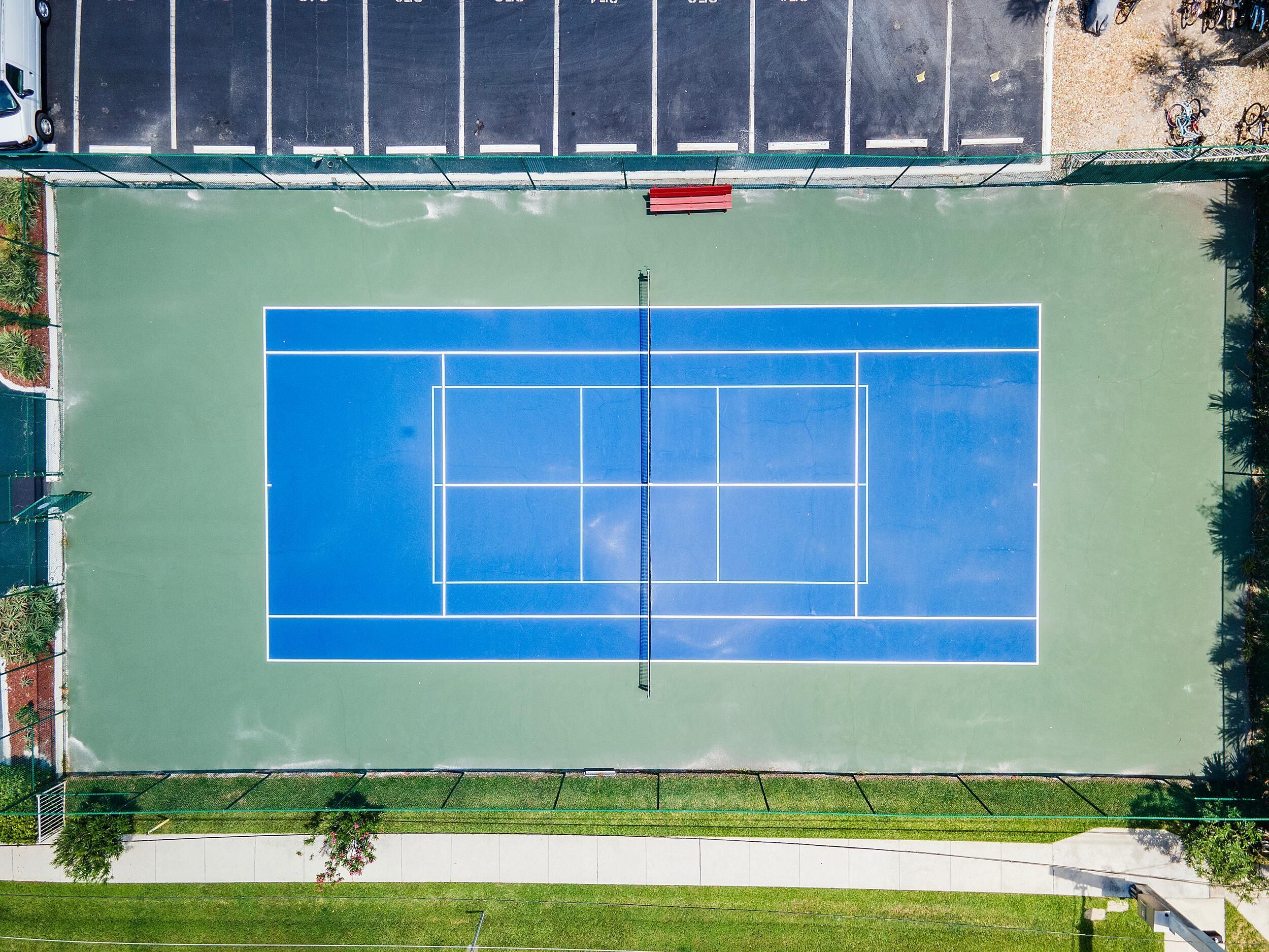 An aerial view of a tennis court with a net, surrounded by a green fence and a parking area with bicycles.