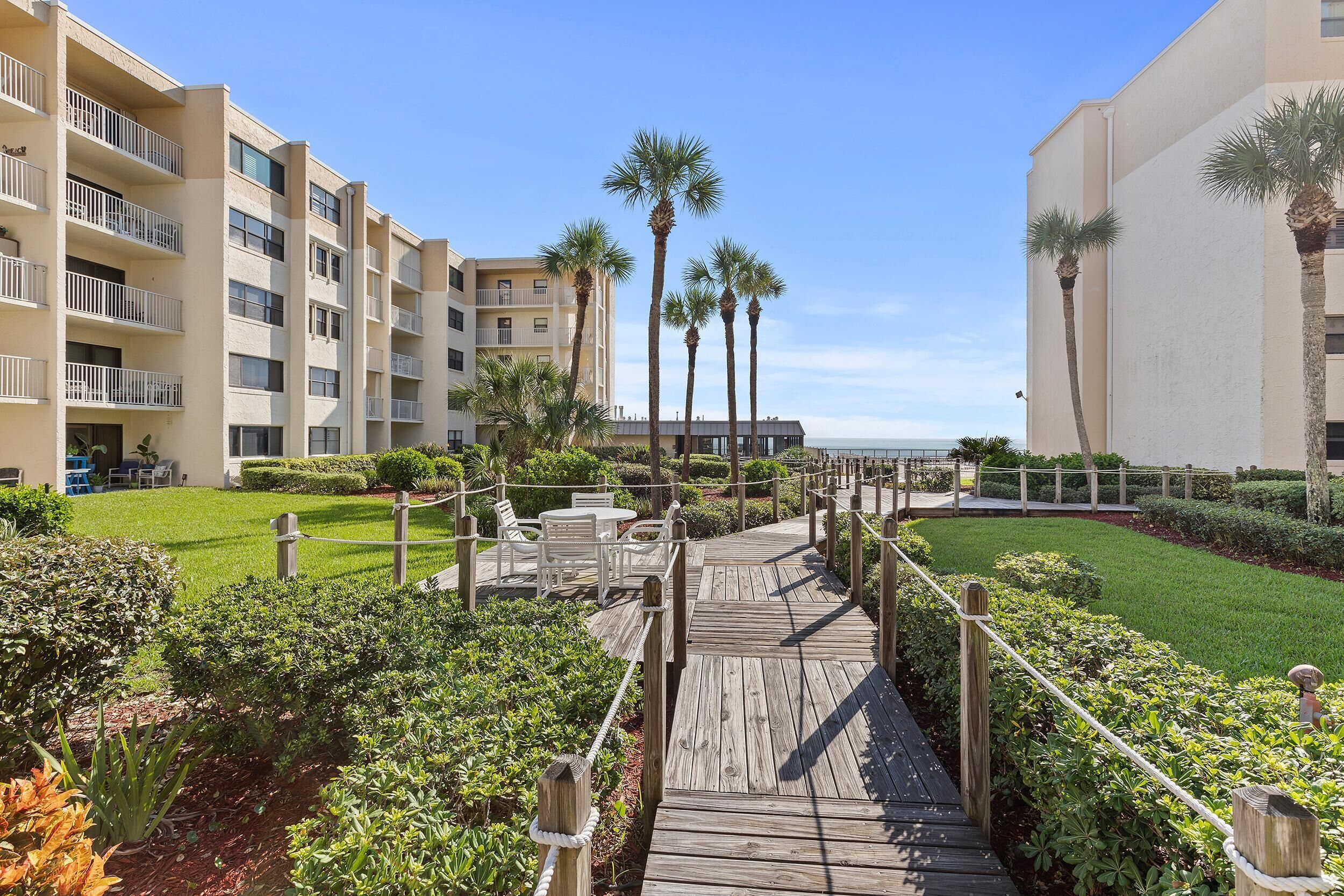 A wooden boardwalk leading to a pool area with palm trees and a view of a hotel building.
