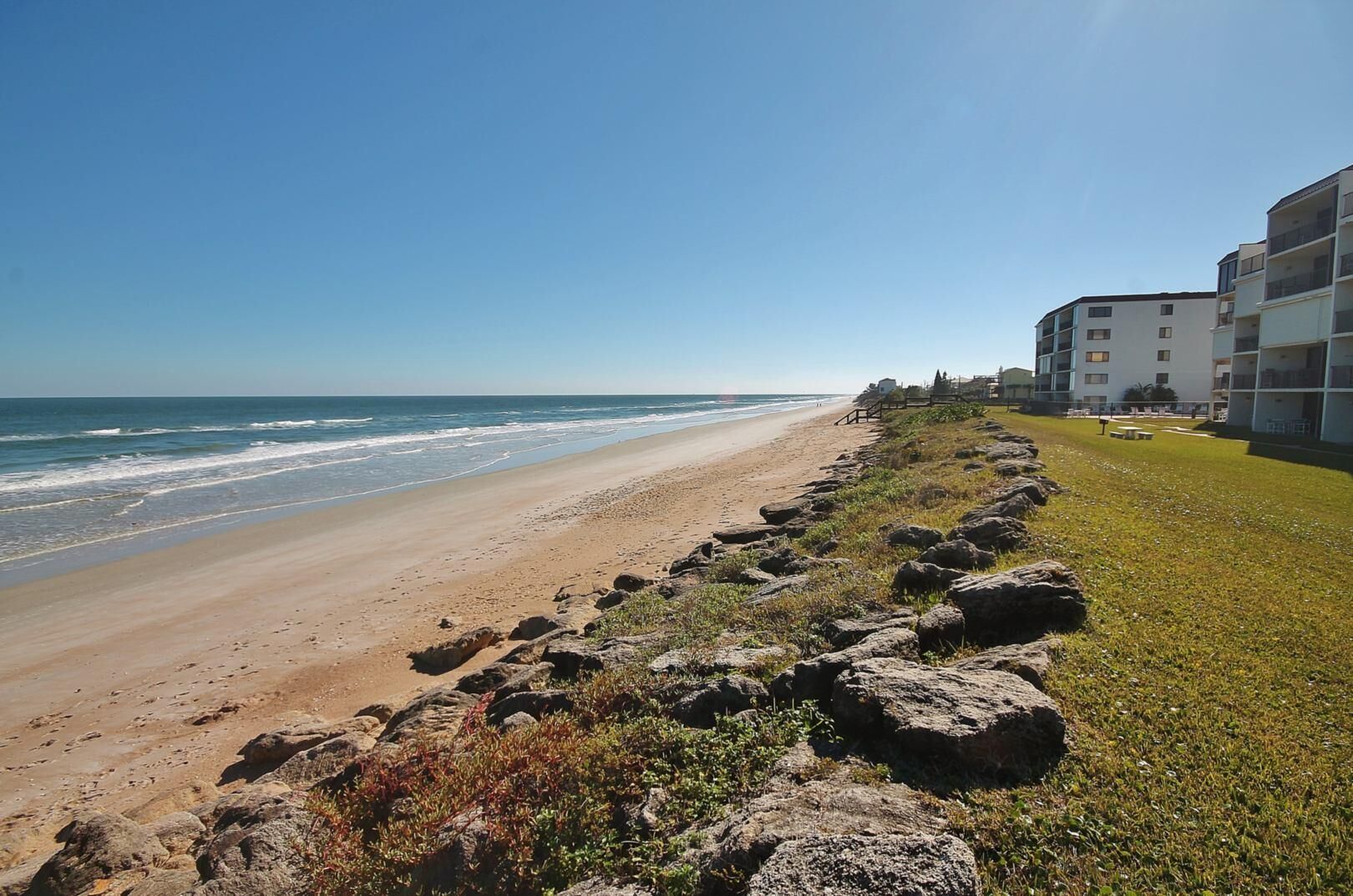 A beach with a rocky shoreline, a grassy area, and a building with multiple floors and balconies.