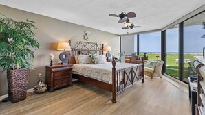 A bedroom with a large bed, bedside lamps, a ceiling fan, a large window with a view of the outdoors, and a wooden floor. - Chadham by the Sea 118 (New Smyrna Beach)