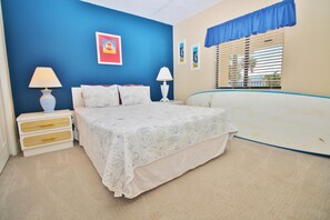 Un dormitorio con una cama, dos mesitas de noche, una mesita auxiliar, una ventana con persianas y un cuadro en la pared.