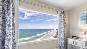 Ein Schlafzimmer mit großem Fenster und Blick auf einen Strand, einem Nachttisch mit Lampe und einem Bett mit floralem Bettzeug.