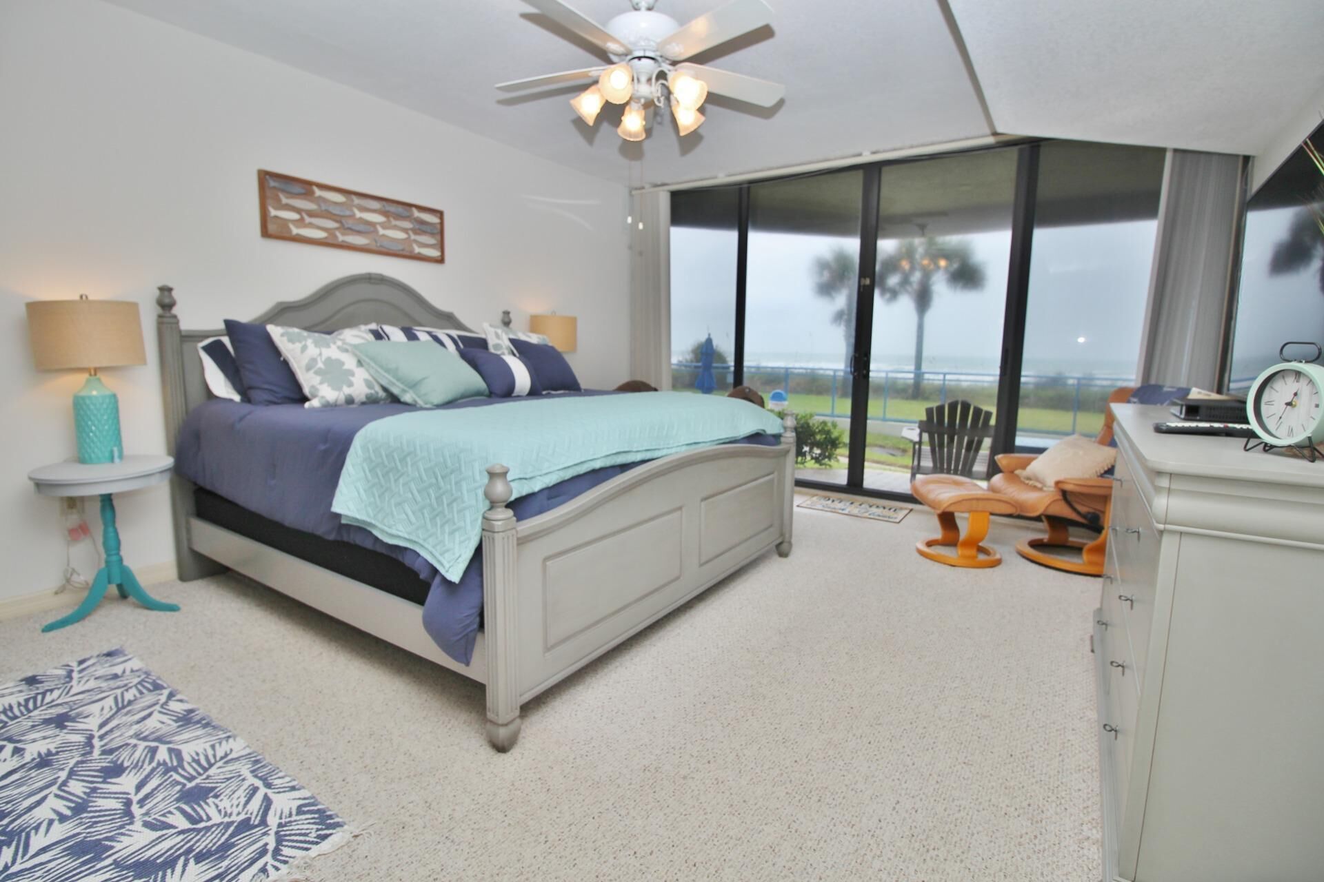 A bedroom with a bed, a nightstand, a lamp, a desk with a chair, and a view of the outdoors.