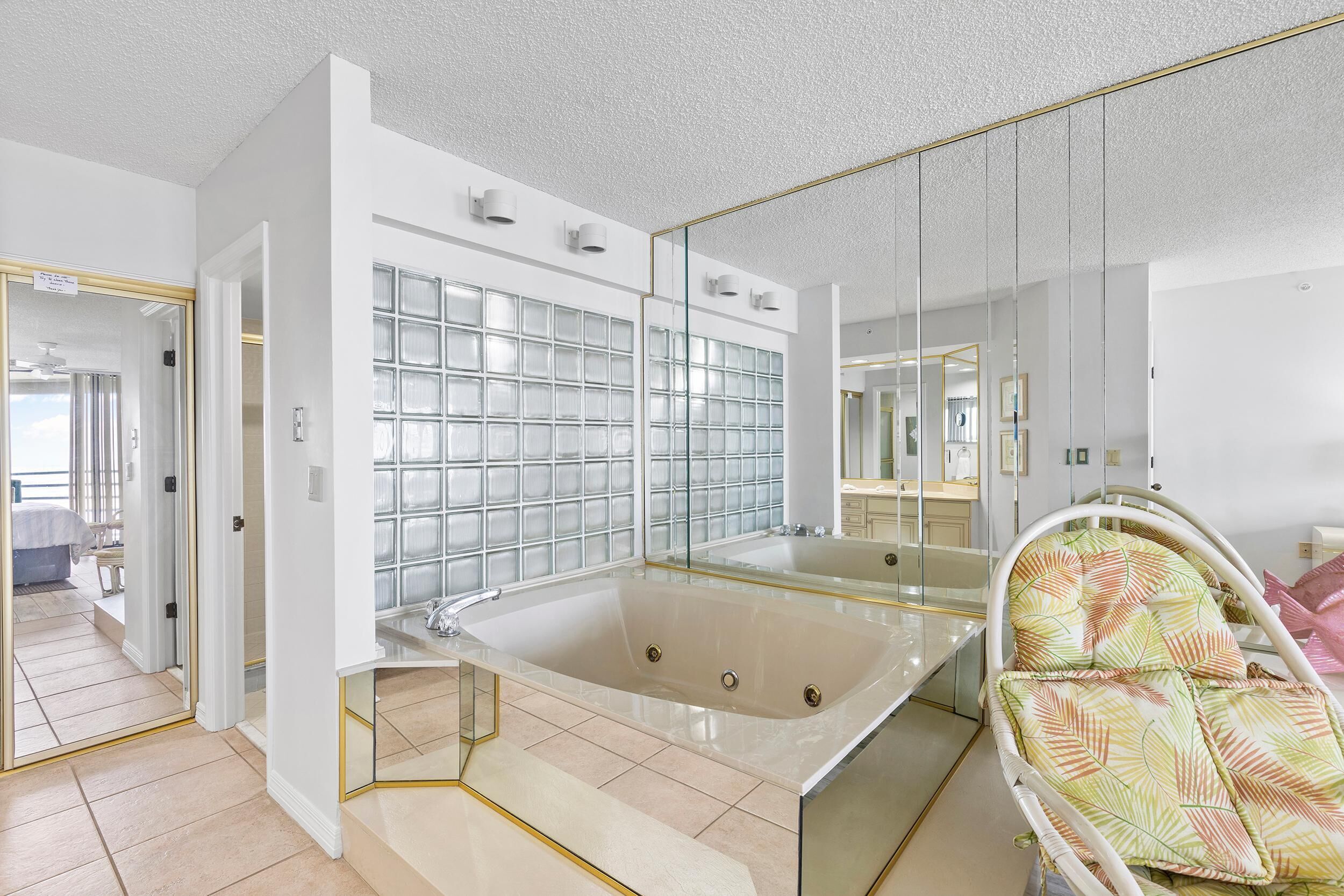 Un baño moderno con bañera de hidromasaje con mampara de vidrio, un espejo grande y una silla de mimbre.