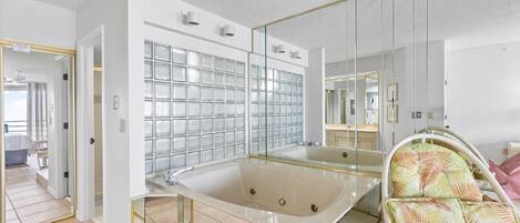 Un baño moderno con bañera de hidromasaje con mampara de vidrio, un espejo grande y una silla de mimbre.