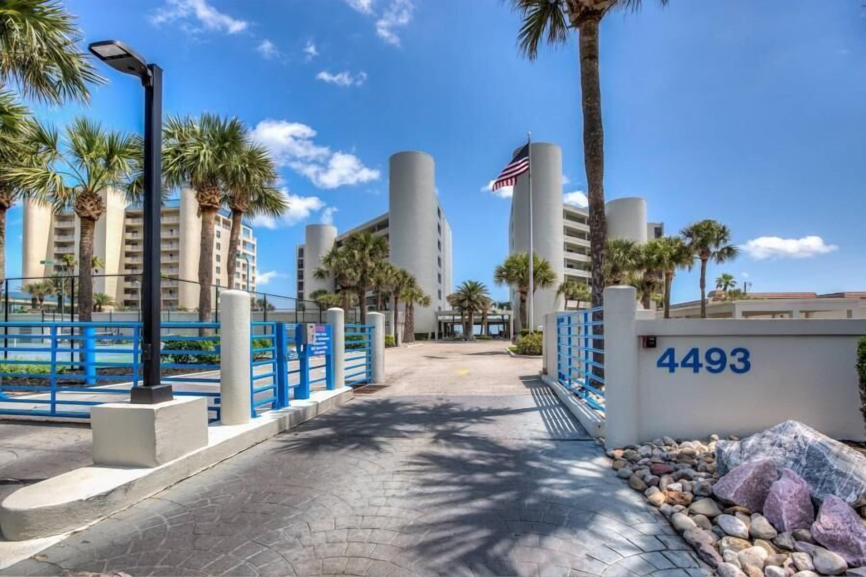 A gated entrance with a blue fence, palm trees, and a building with the number 4493.