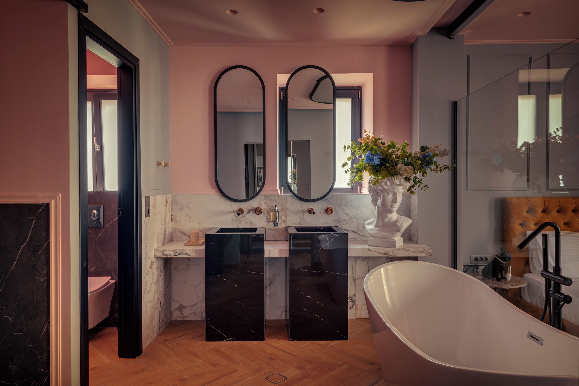 Un baño moderno con una bañera independiente, encimeras de mármol, dos espejos grandes y un jarrón con flores.