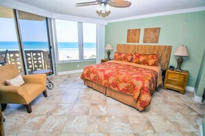 A bedroom with a large bed, a chair, a nightstand, and a view of the beach. - Golden Arms 503 (New Smyrna Beach)