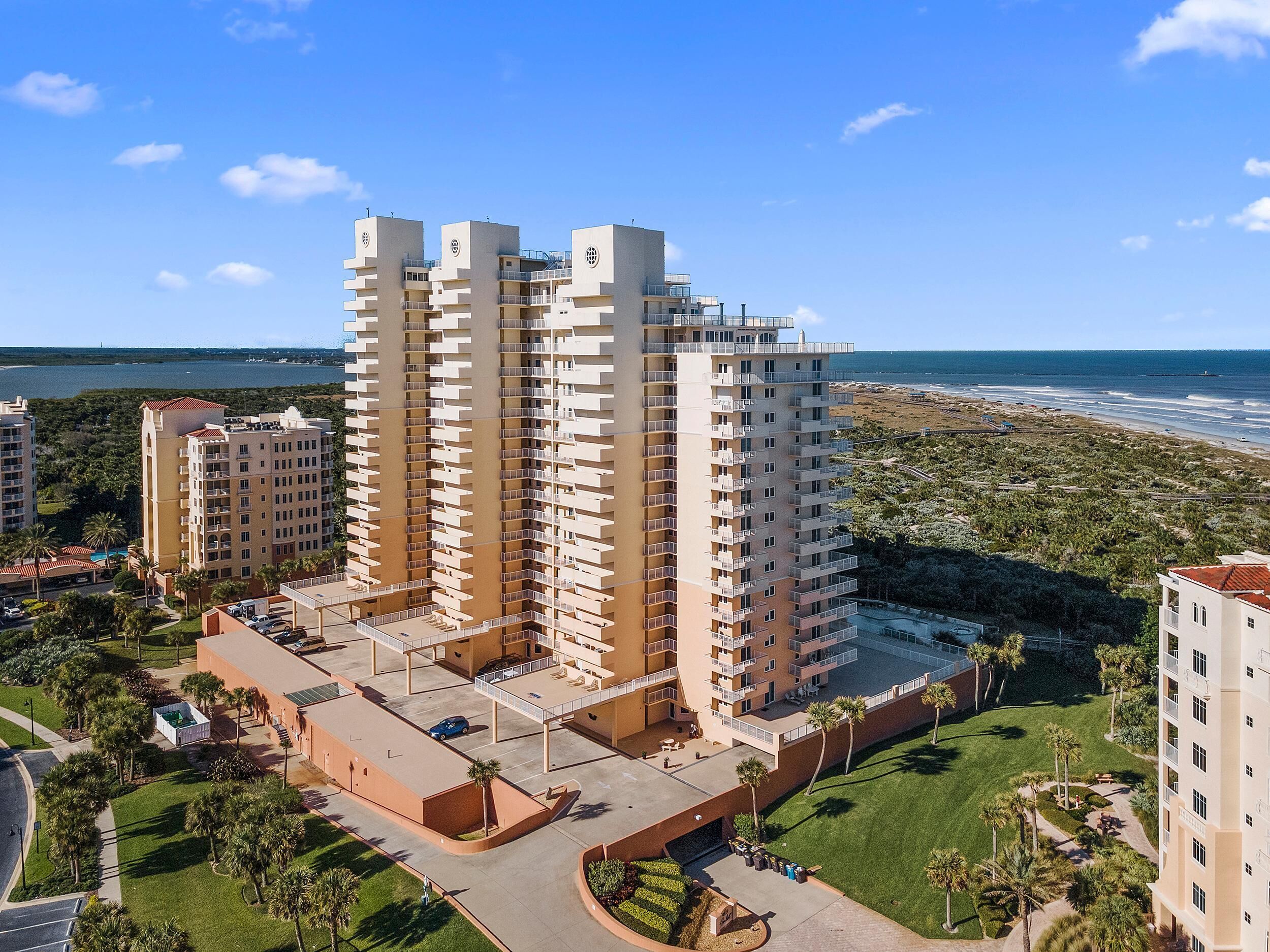 A high-rise hotel with multiple balconies, a parking area, and a view of the beach.