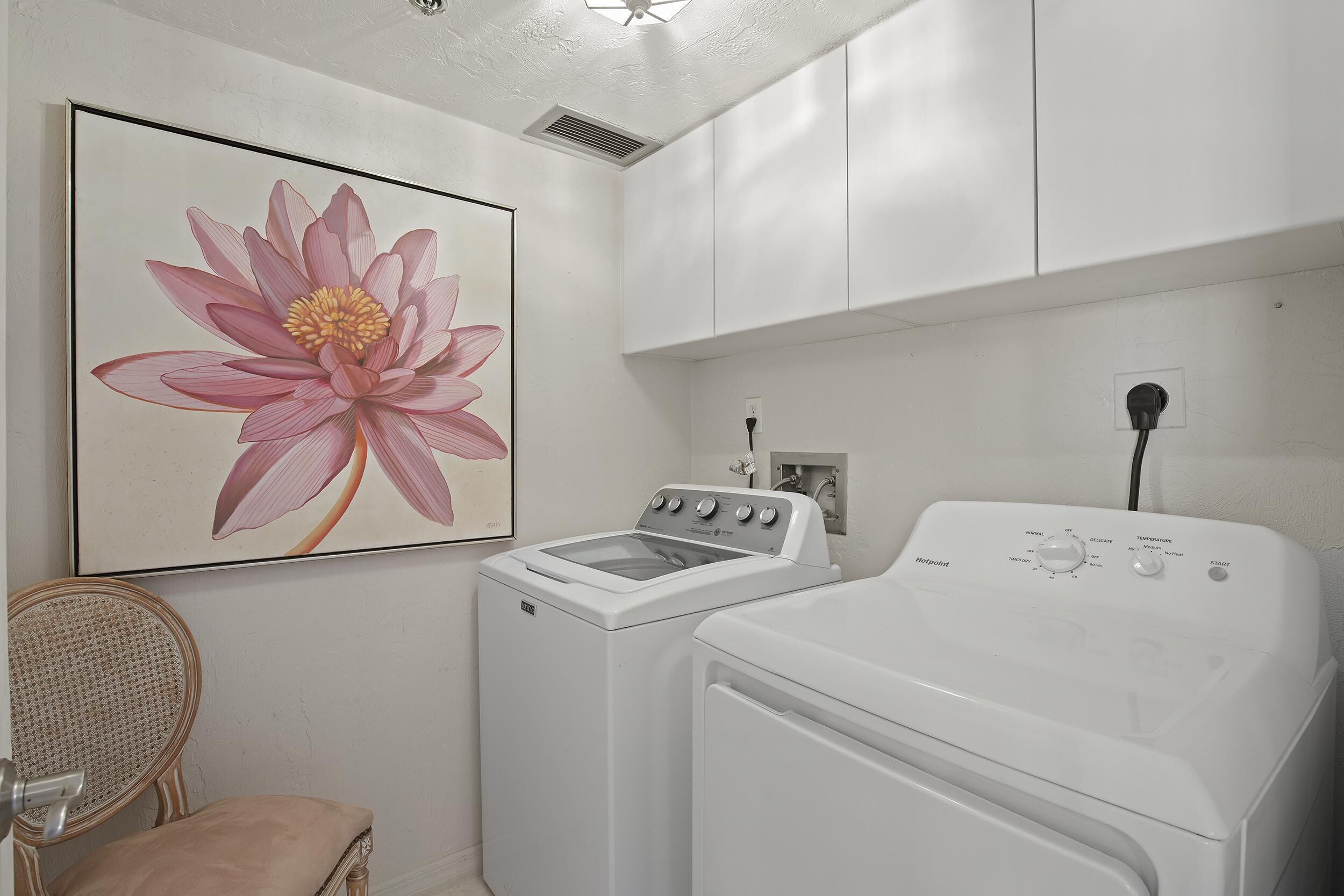 A laundry room with a washing machine and dryer, a wicker chair, and a large floral painting on the wall.