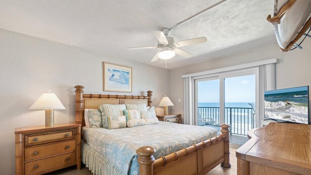 A bedroom with a wooden bed, two bedside tables with lamps, a ceiling fan, a large window with a view of the ocean, and a painting on the wall. - Hacienda del Sol II 413 (New Smyrna Beach)