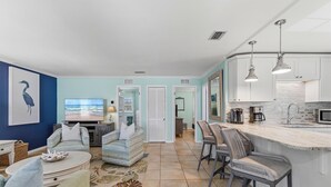 A spacious living area with a large TV, comfortable seating, a kitchen with white cabinets, and a tiled floor.