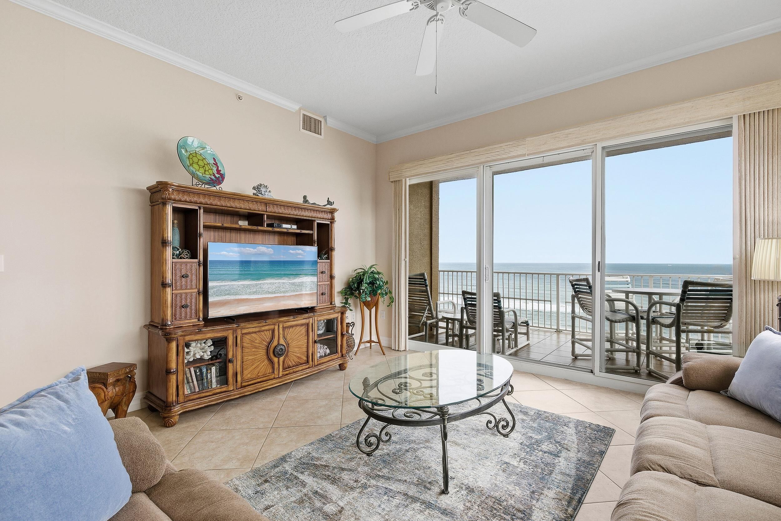 A living room with a glass coffee table, a sofa, a TV on a wooden cabinet, and a balcony with a view of the ocean.