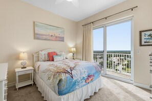 A bedroom with a large bed, a nightstand, a lamp, a painting, and a view of the outside. - Malibu 704 (New Smyrna Beach)