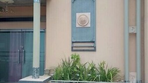 A building facade with a beige wall, a blue window with a circular design, and a small garden area with plants. - Upstairs Luxury (Lusaka)