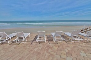 Une plage avec des chaises longues blanches alignées sur une zone pavée, face à l’océan.