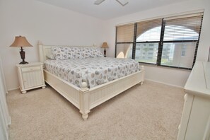 Une chambre à coucher comprenant un lit, deux tables de chevet, une commode, une fenêtre avec stores et un ventilateur de plafond.