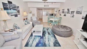 A modern living room with a sofa, a coffee table, a bean bag, and a TV. There is a kitchen area in the background with a dining table and chairs. - Ocean Club North C208 (New Smyrna Beach)