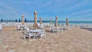 An outdoor seating area with white metal chairs and tables, beige umbrellas, and a view of the ocean. - Ocean Club North C208 (New Smyrna Beach)