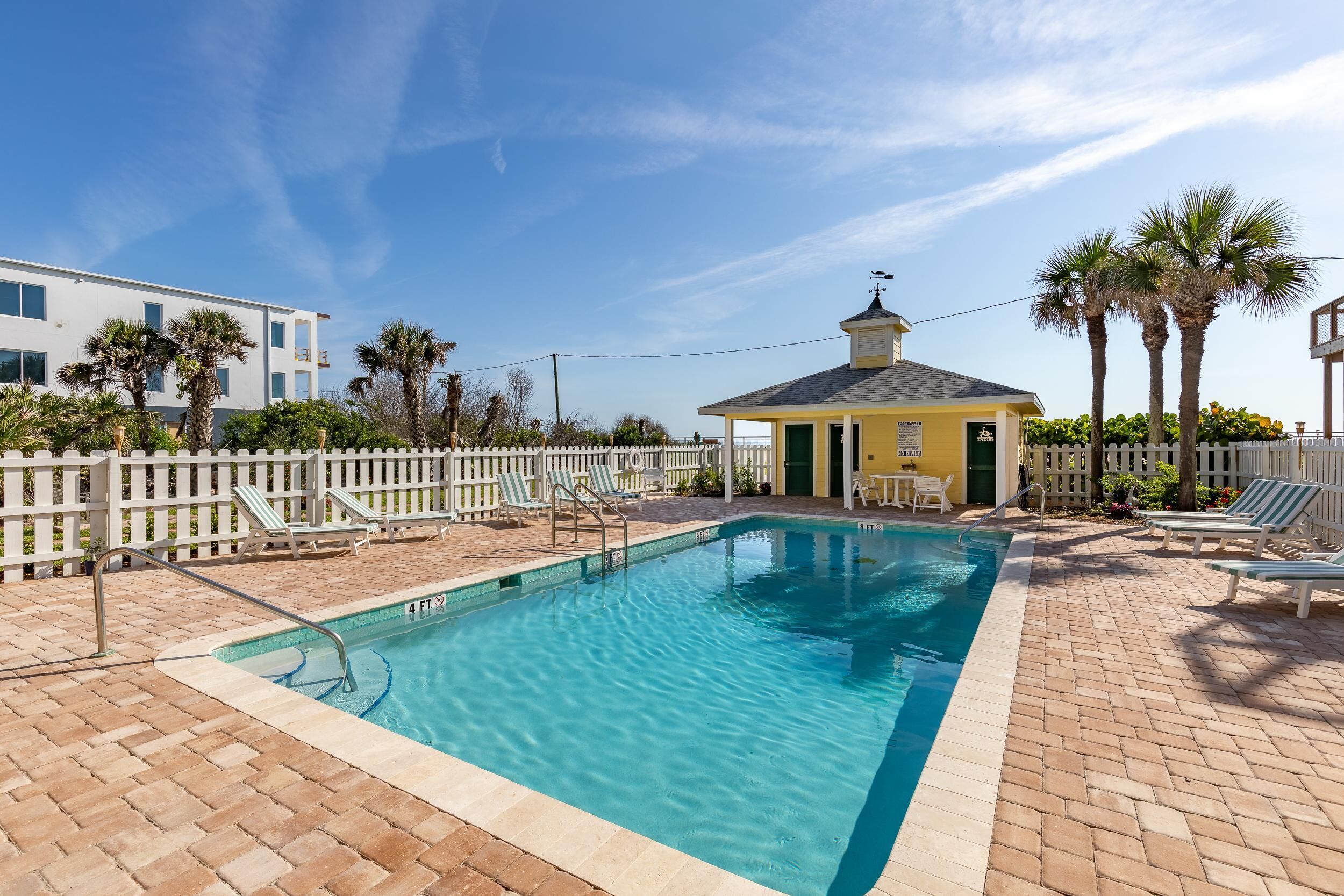 A rectangular pool with a 4 FT depth marker, surrounded by a white fence, palm trees, and a small yellow building with a red roof.