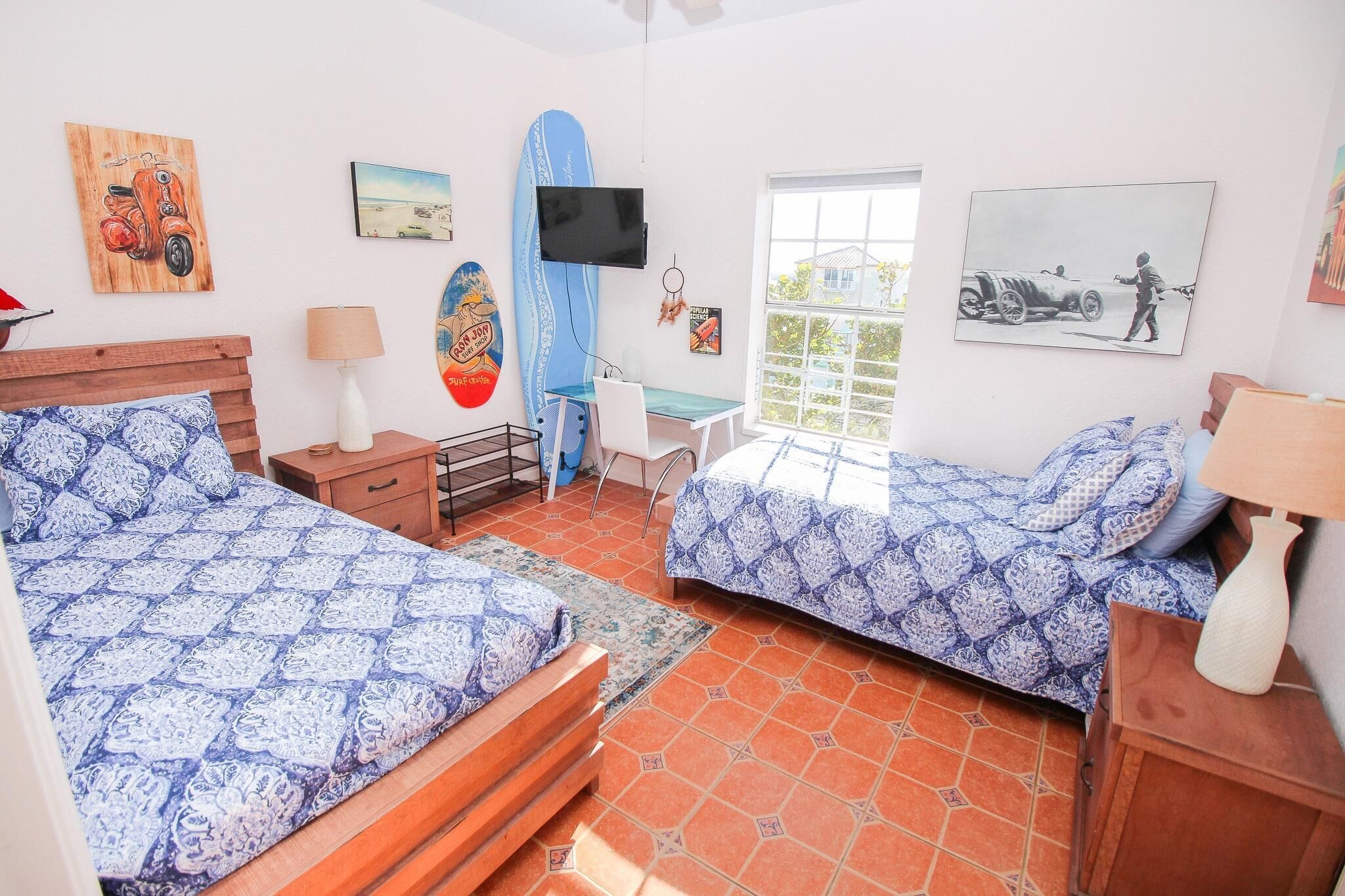 A bedroom with two beds, a desk, a chair, a TV, and artwork on the walls.