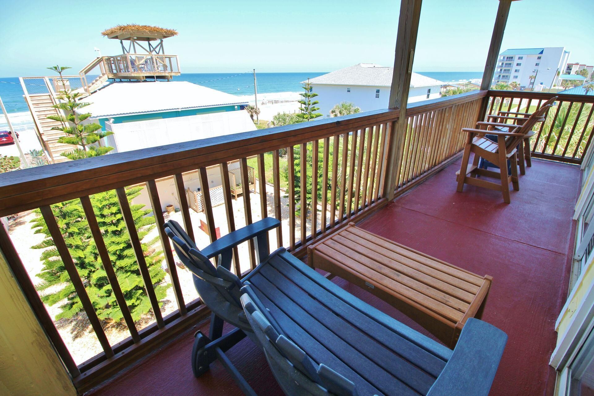 A balcony with wooden furniture, a view of the beach, and a building in the background.