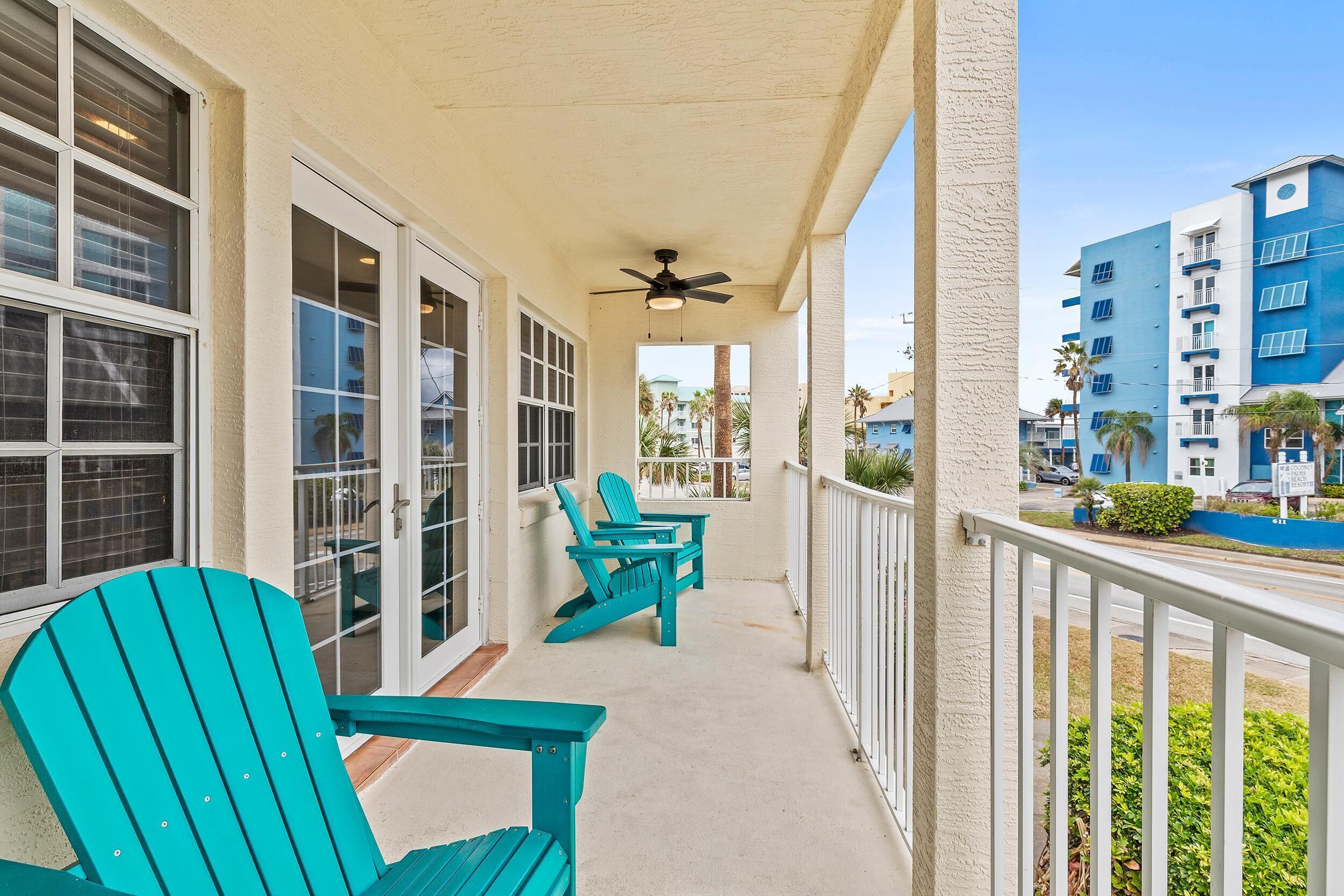A balcony with teal Adirondack chairs, a ceiling fan, and a view of a blue building.