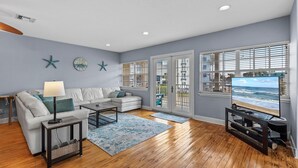 A living room with a white sofa, a coffee table, a TV stand, and a large window with a view of the beach. - Ocean's Edge (New Smyrna Beach)