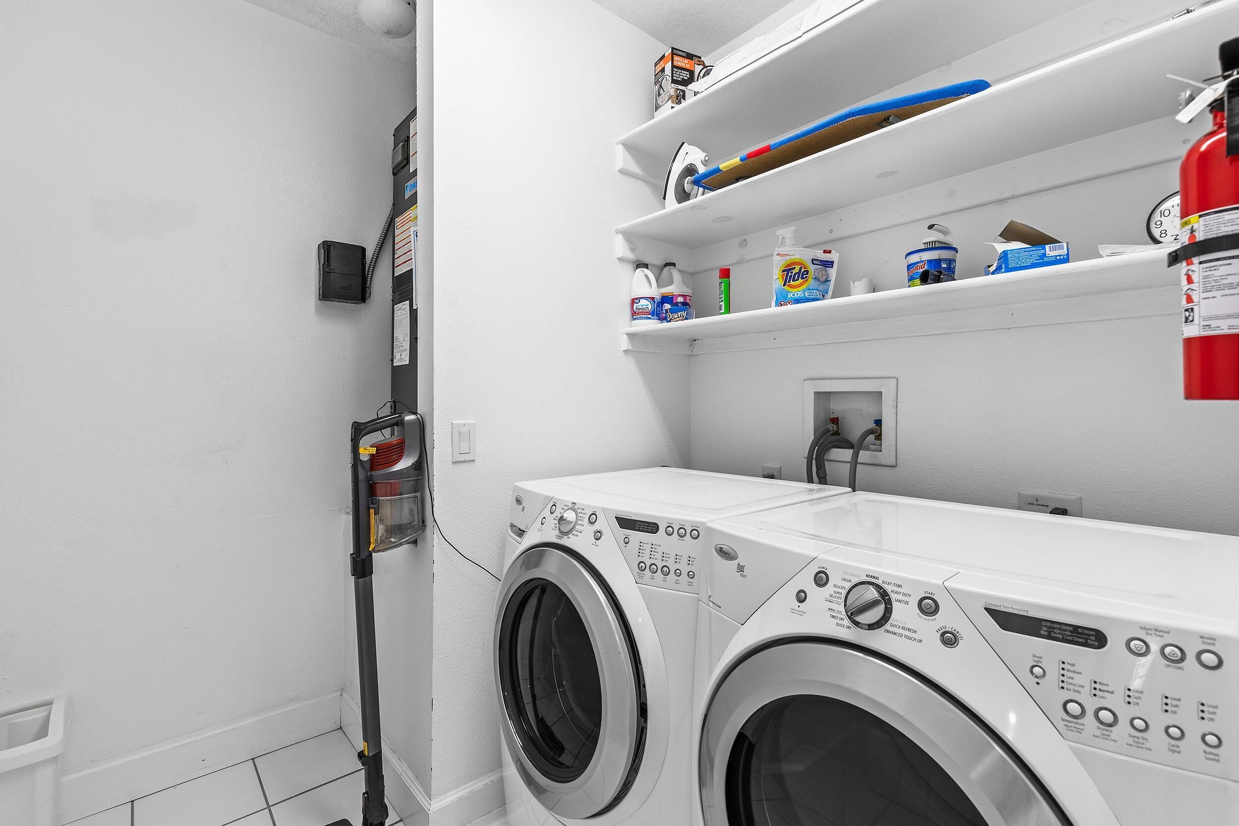Een wasruimte met een wasmachine, planken met schoonmaakmiddelen en een brandblusser.