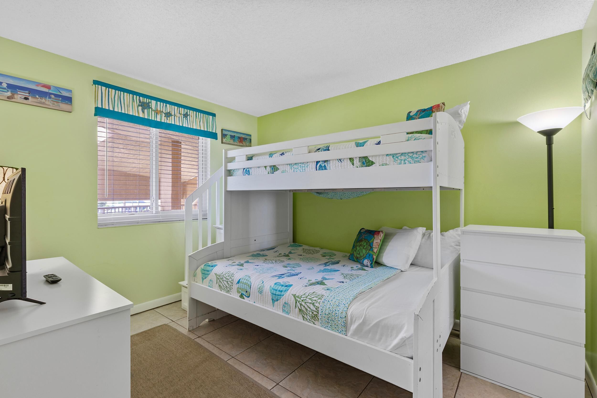 Une chambre avec un lit superposé, un bureau et une commode, des murs verts et une fenêtre avec des stores.