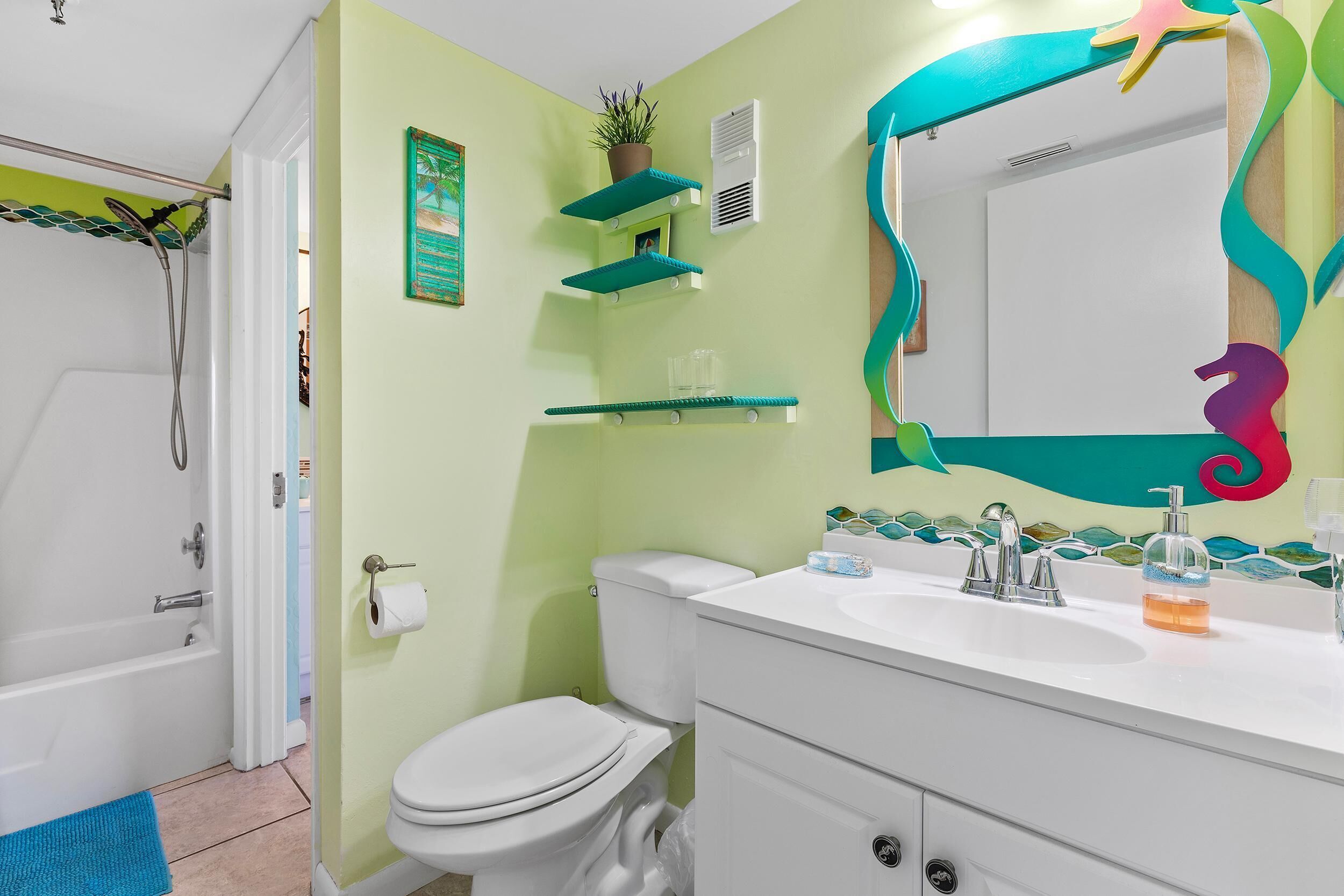 A bathroom with a white toilet, a white sink, and a mirror with a colorful frame. There are shelves above the toilet and a shower curtain rod.