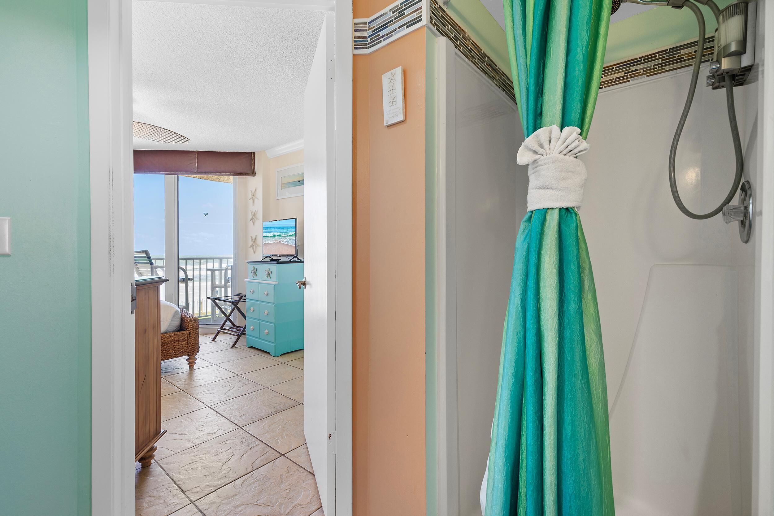 A bathroom with a shower curtain, a towel hanging on the door, and a view of the beach through the window.