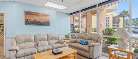 Amplia sala de estar con dos sofás color beige, una mesa de centro de madera y una mesita auxiliar con una lámpara. Un ventanal ofrece vistas a un edificio y palmeras.