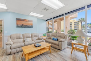 A spacious living area with two beige sofas, a wooden coffee table, and a side table with a lamp. A large window offers a view of a building and palm trees. - Oceania Plaza 405 (New Smyrna Beach)