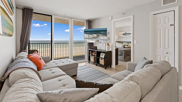 A living room with a large window overlooking a beach, a sofa, a coffee table, and a bookshelf. - Oceania Plaza 405 (New Smyrna Beach)