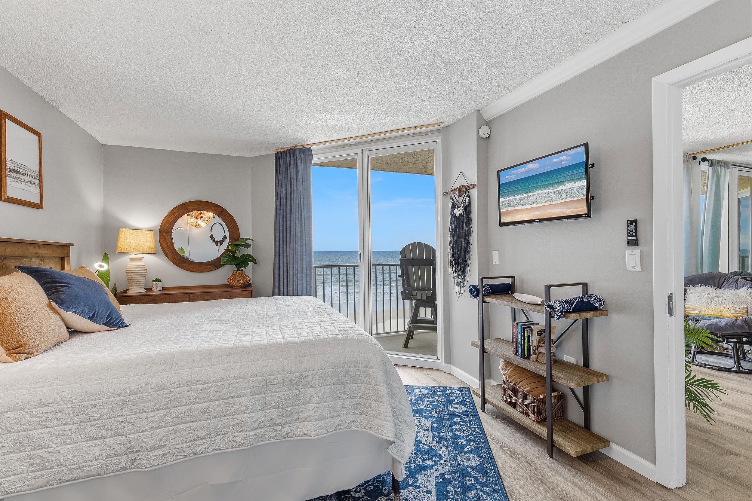 Un dormitorio con cama, escritorio, silla, televisor y balcón con vista al mar.